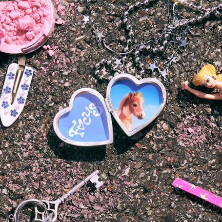1st EP focus by hearts2hearts cover art, a photo of things fallen on the ground such as a heart chaped locket necklace that has the title of the album written in it with a photo of a pony in the other half, other things are pink blush, white hairclip, a doll, a key, and silver glitter that is star shaped